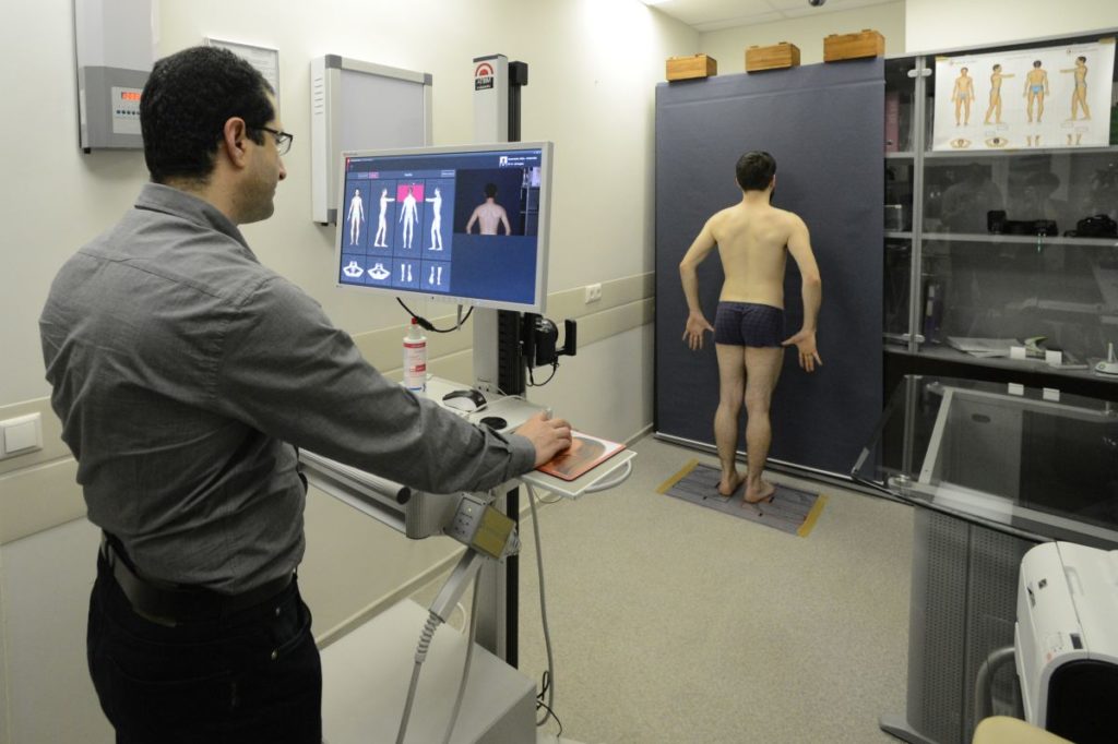 Процесс съемки при помощи аппарата FotoFinder Bodystudio ATBM. Пациент стоит на коврике перед специальным фоном. На мониторе доктор видит, какое положение тела в данный момент должен занять пациент для съемки, перед монитором расположена камера, которая двигается по вертикали