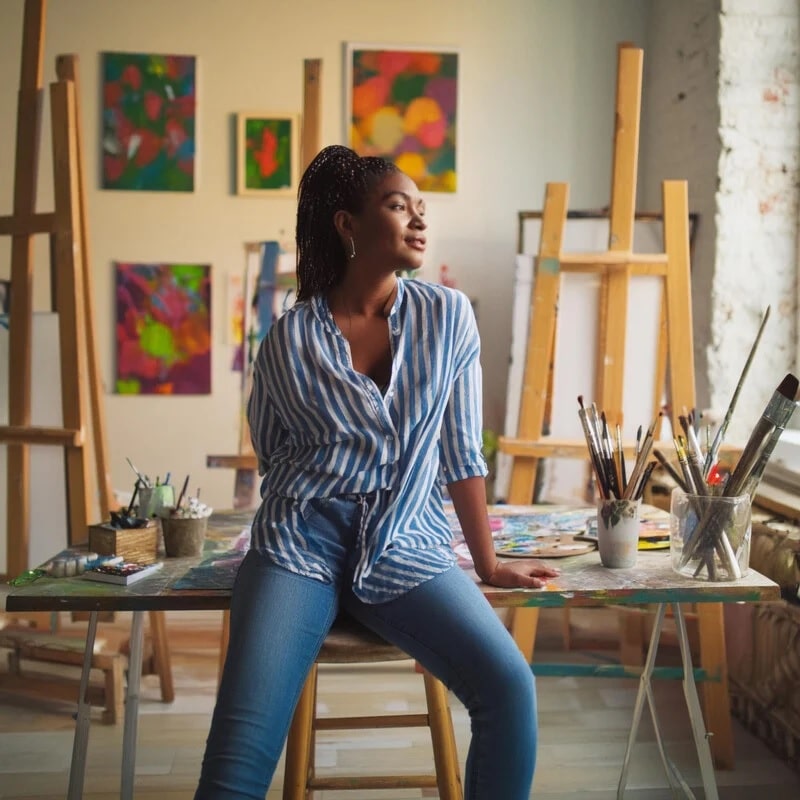  Сгенерированное изображение по подсказке: художница в своей студии сидит на столе и выглядит задумчивой среди множества картин / © Adobe